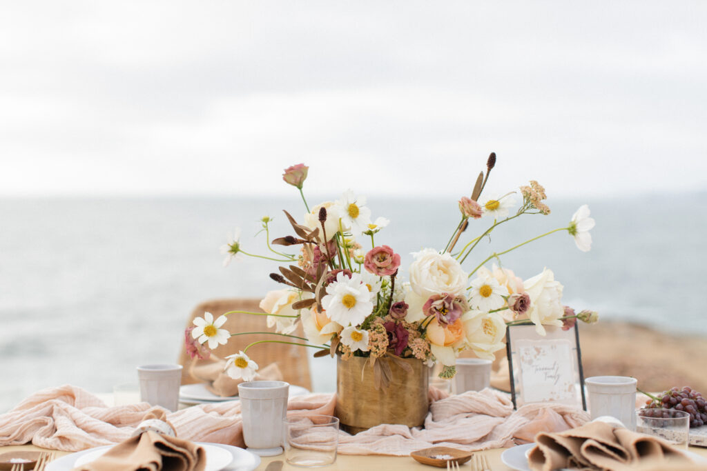 Elegant outdoor dining setup on a seaside cliff featuring a floral centerpiece by a Charlotte wedding florist, draped linens, and a spread of grapes and glasses.