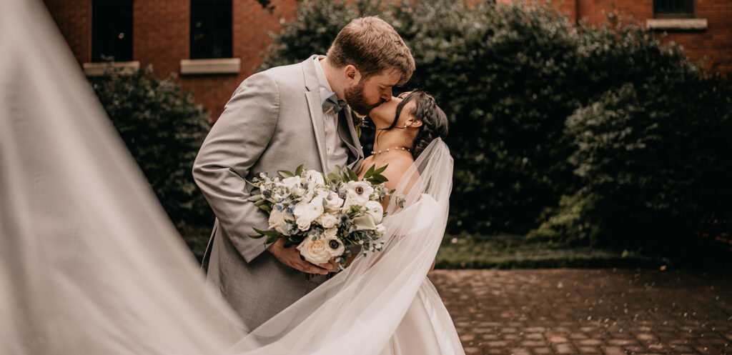 A wedding couple shares a kiss outdoors, the bride holding a bouquet designed by a Charlotte florist and her veil flowing.