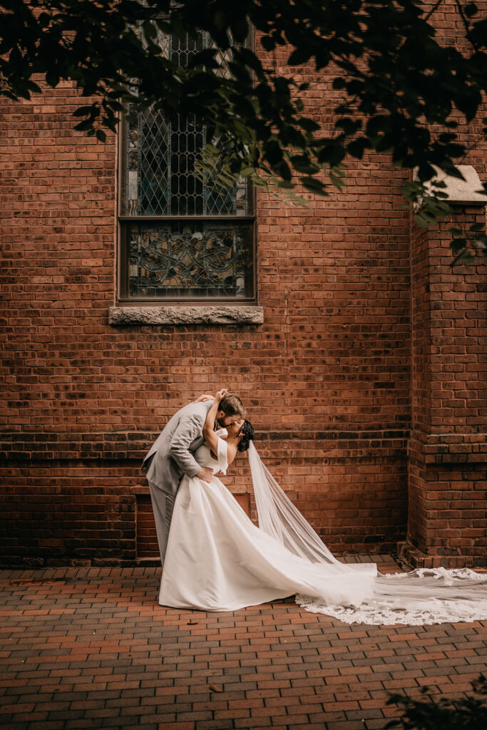A bride and groom kiss tenderly outside a brick building with stained glass windows, framed by green leaves arranged by a Charlotte florist.
