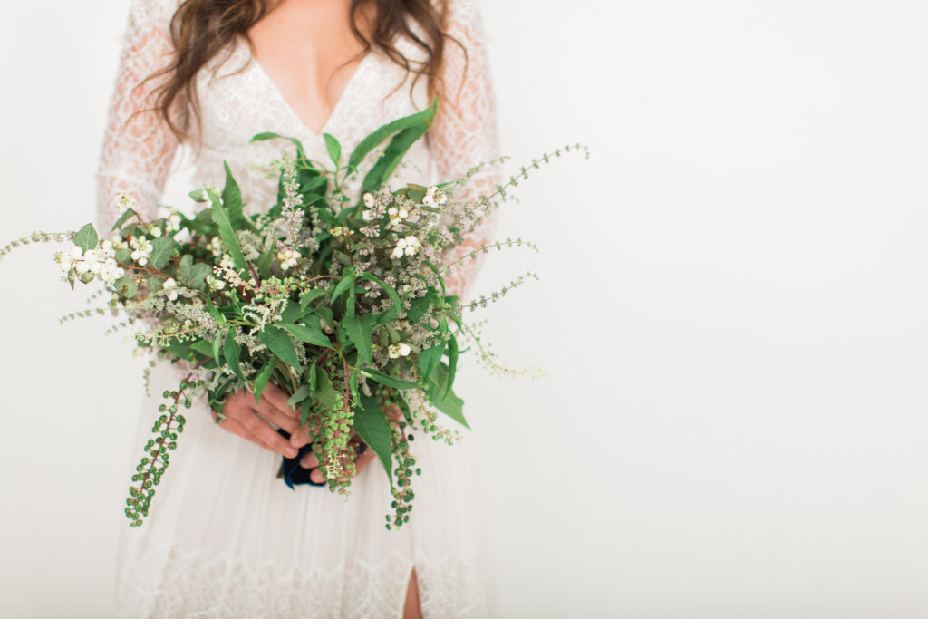 A bride in a lace wedding dress arranged by a wedding planner in Charlotte holds a large, green and white bouquet crafted by a Charlotte wedding florist, with only her torso visible.