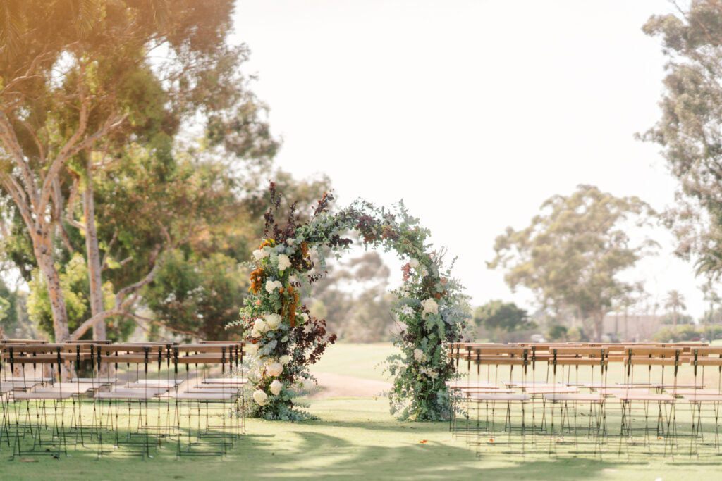 An ornate floral archway stands at the end of an aisle lined with wooden chairs, set in a sunlit outdoor area with trees in the background.