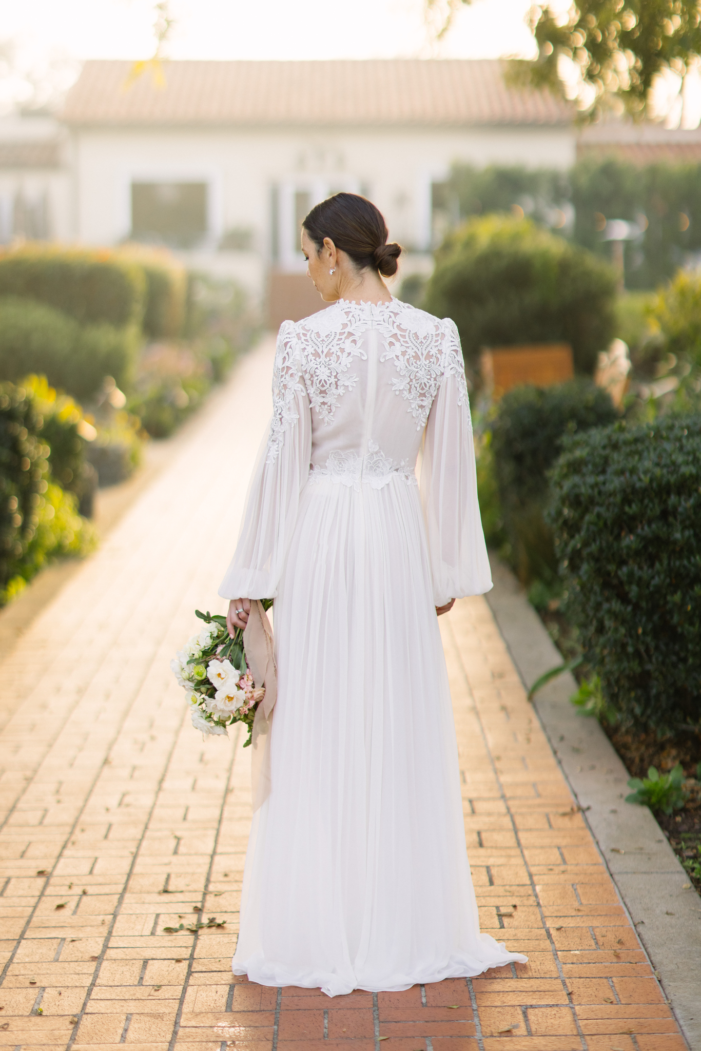 Bride in a white lace gown holding a bouquet crafted by a Charlotte wedding florist, walking away on a brick pathway, surrounded by greenery.