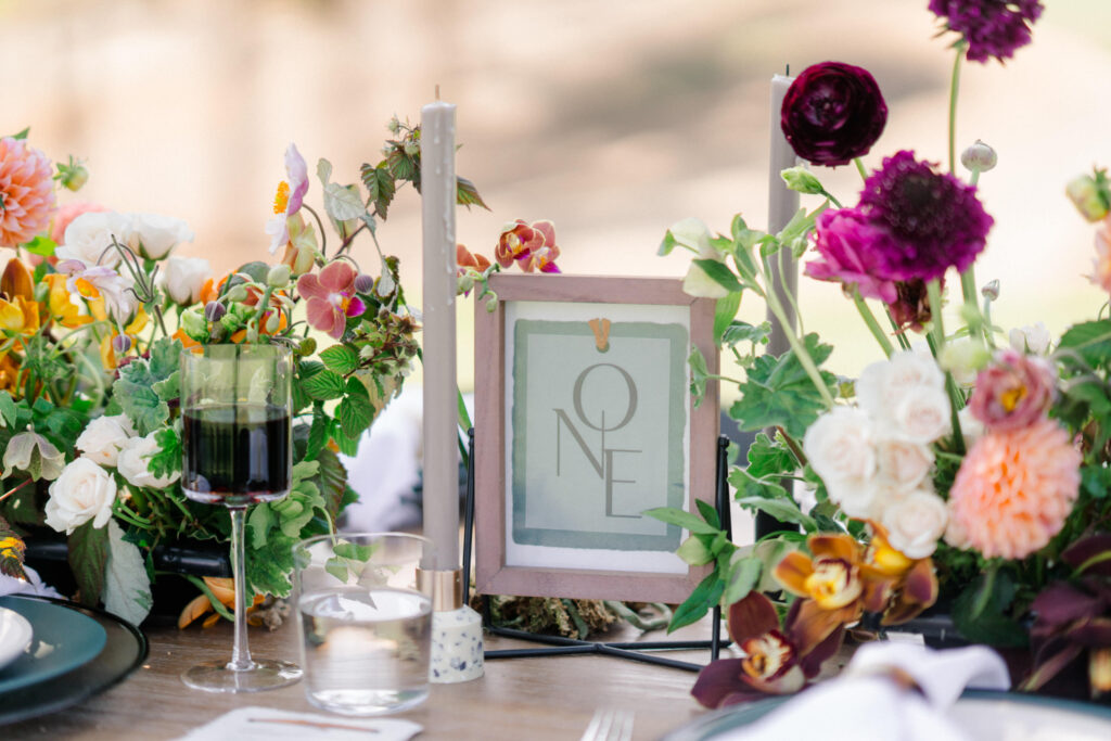 Elegant outdoor dining table setup with a floral centerpiece by top wedding florists in Charlotte NC, a framed table number, wine glasses, and candles, conveying a sophisticated ambiance.