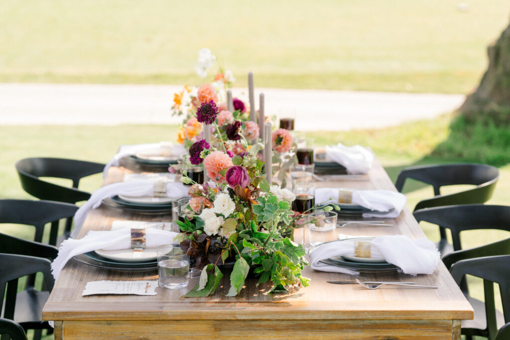 Elegant outdoor dining setup with a wooden table adorned by a floral centerpiece from a Charlotte wedding florist, set with plates, glasses, and black chairs on a grassy field.