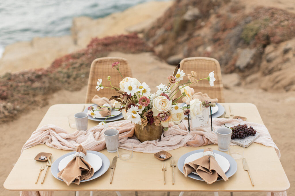 Elegant beachside dining setup featuring a table with a floral centerpiece designed by a Charlotte wedding florist, plates, and soft linens, overlooking a cliff.