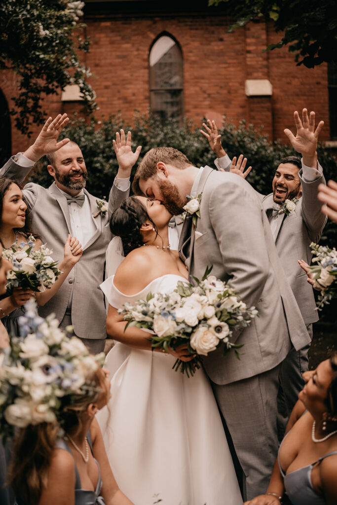 A bride and groom kiss tenderly outside a brick building with stained glass windows, framed by green leaves arranged by a Charlotte florist.