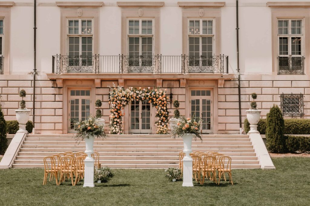 grand wedding venue estate with large stairs- ceremony site on steps and lawn with wooden chairs, large urn entry floral pieces, large floral arch at altar in colors of peach and neutrals