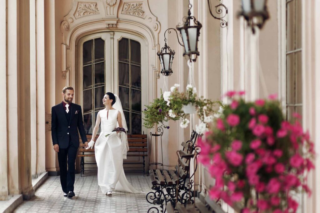 bride and groom holding hands and walking with large luxury estate as backdrop- moody aesthetic attire