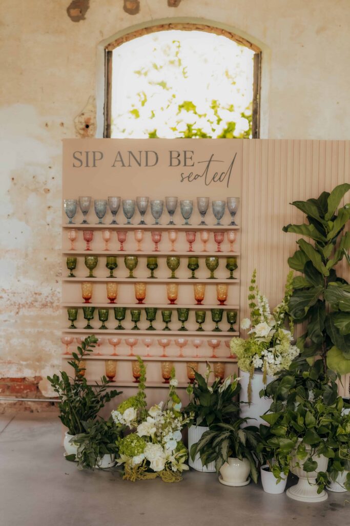 Sip and be seated luxury wedding backdrop installation with colored vintage glassware at Providence Cotton Mill in Maiden, NC