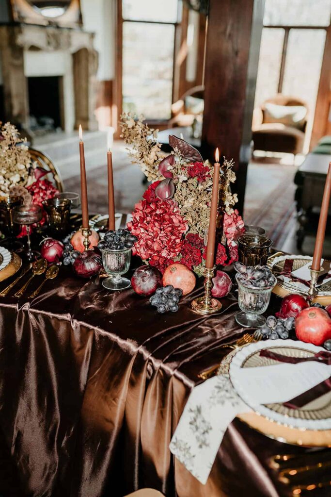 Sunset Ridge Estate luxury southern wedding venue in Monroe NC with moody winter reception table in front of windows at large estate with gold candles and large florals and fruit in shades of chocolate brown and burgundy by Crafted Charm Florals