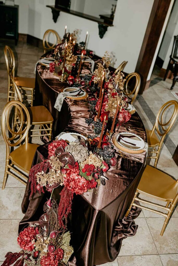 Sunset Ridge Estate luxury southern wedding venue in Monroe NC with moody winter reception table in front of windows at large estate with gold candles and large florals and fruit in shades of chocolate brown and burgundy by Crafted Charm Florals