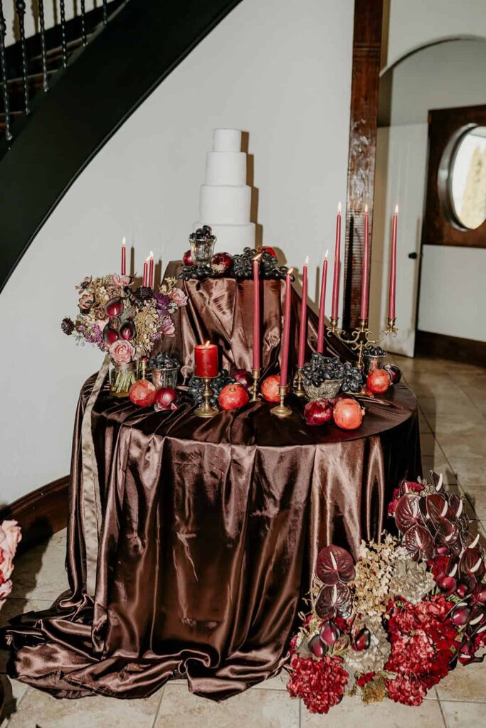 Sunset Ridge Estate luxury southern wedding venue in Monroe NC with moody winter cake table with draped linens in front of stairs at large estate with gold candles and large florals and fruit in shades of chocolate brown and burgundy by Crafted Charm Florals