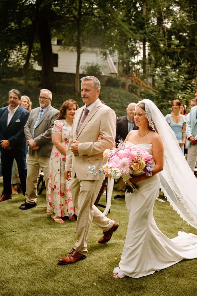 Father walking bride during ceremony carrying large floral bouquet of spring colors and peonies at the Historic Robertson Homestead in Rock Hill, SC